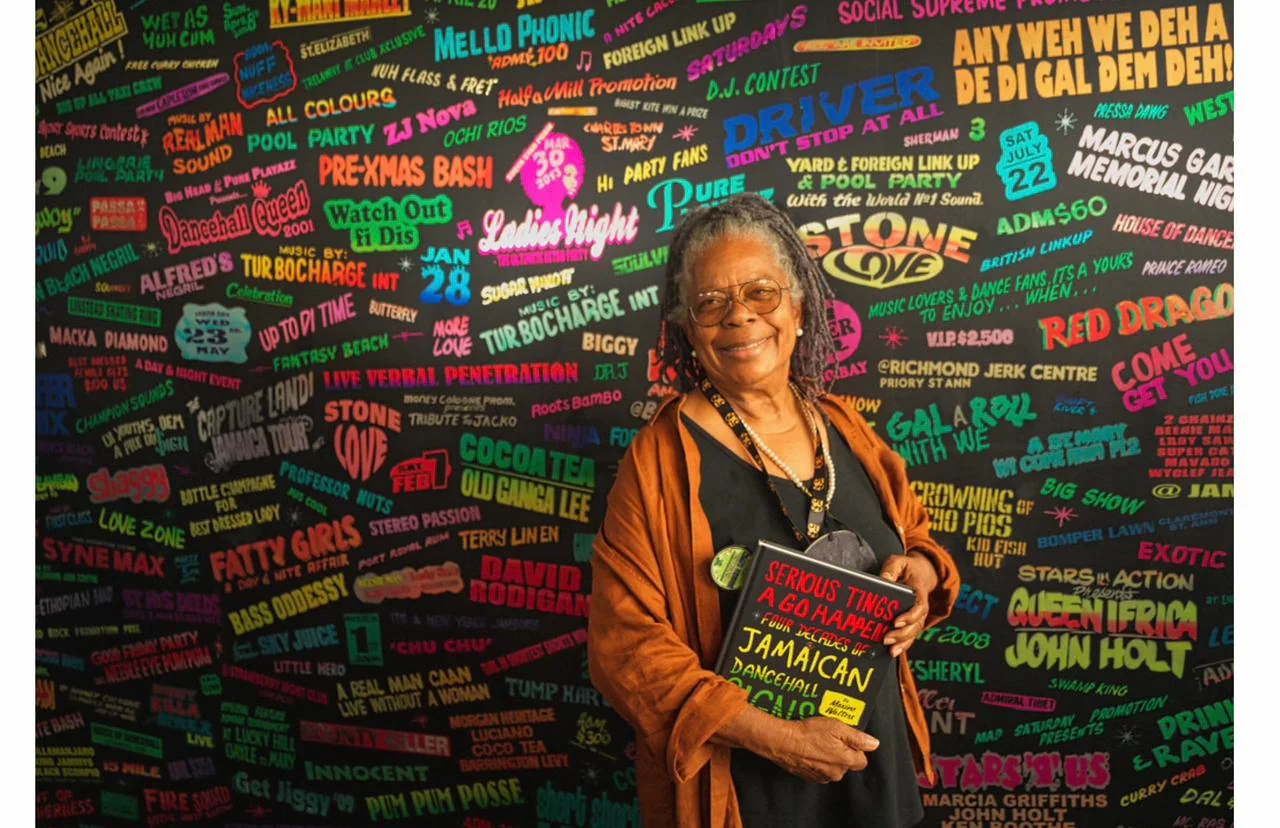 Maxine Walters with her book in front of a wall of hand-painted Jamaican dancehall posters